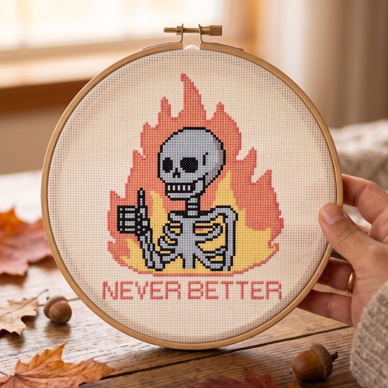 May include: A circular wooden embroidery hoop containing a cross-stitch design of a light blue skeleton giving a thumbs-up gesture, surrounded by orange and yellow flames, with the text NEVER BETTER stitched in red letters below the skeleton.