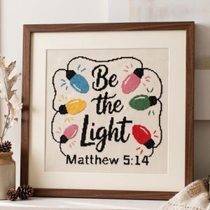 Może przedstawiać: Oprawiony haft krzyżykowy na kremowej tkaninie z czarnym napisem Be the Light Matthew 5:14 otoczony sznurem kolorowych lampek choinkowych w kolorze niebieskim, ż&oacute;łtym, r&oacute;żowym, zielonym i czerwonym, w ciemnobrązowej drewnianej ramie.