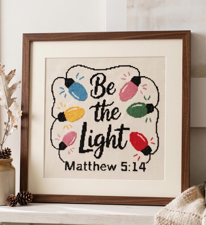 Może przedstawiać: Oprawiony haft krzyżykowy na kremowej tkaninie z czarnym napisem Be the Light Matthew 5:14 otoczony sznurem kolorowych lampek choinkowych w kolorze niebieskim, ż&oacute;łtym, r&oacute;żowym, zielonym i czerwonym, w ciemnobrązowej drewnianej ramie.