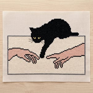 Pode incluir: Um bordado em ponto cruz sobre tecido Aida de cor creme, apresentando um gato preto estendendo uma pata em dire&ccedil;&atilde;o a uma m&atilde;o humana. O design &eacute; emoldurado por uma fina borda retangular cinza, com o gato e as m&atilde;os em estilo pixelizado.