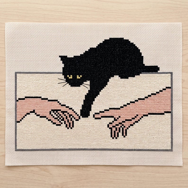 Pode incluir: Um bordado em ponto cruz sobre tecido Aida de cor creme, apresentando um gato preto estendendo uma pata em dire&ccedil;&atilde;o a uma m&atilde;o humana. O design &eacute; emoldurado por uma fina borda retangular cinza, com o gato e as m&atilde;os em estilo pixelizado.