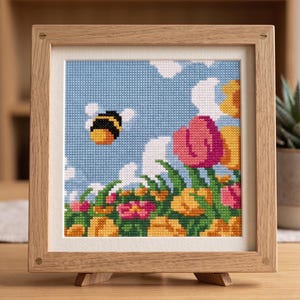 K&ouml;nnte beinhalten: Kreuzstich-Kunstwerk einer Biene, die &uuml;ber einem Blumenfeld fliegt. Das Kunstwerk zeigt einen blauen Himmel mit wei&szlig;en Wolken und farbenfrohen Blumen in Rosa, Gelb und Gr&uuml;n. Das gerahmte St&uuml;ck wird auf einem Holzst&auml;nder pr&auml;sentiert.