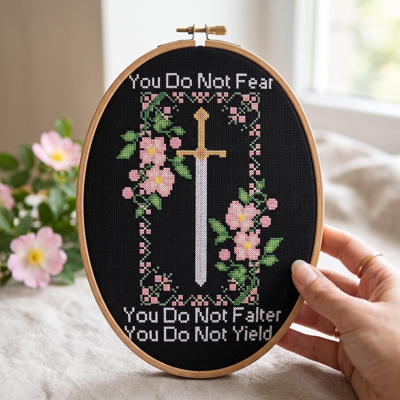 May include: An oval wooden embroidery hoop containing black fabric with a cross-stitched design. The design features a central sword surrounded by pink flowers and green leaves, with the text "You Do Not Fear, You Do Not Falter, You Do Not Yield."