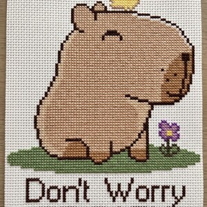 May include: A cross-stitch embroidery on white fabric featuring a tan capybara with a small yellow bird on its head, standing on a patch of green grass next to a purple flower. The text below reads Don't Worry BE CAPY in dark brown thread.