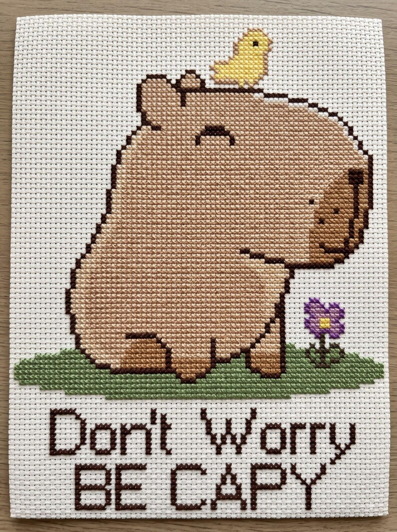 May include: A cross-stitch embroidery on white fabric featuring a tan capybara with a small yellow bird on its head, standing on a patch of green grass next to a purple flower. The text below reads Don't Worry BE CAPY in dark brown thread.