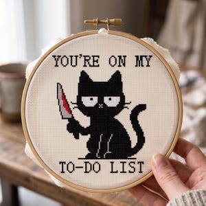 Pu&ograve; includere: Un telaio da ricamo circolare in legno contenente tessuto bianco con un disegno a punto croce di un gatto nero che tiene in mano un coltello. Il testo sopra il gatto recita YOU'RE ON MY e il testo sotto TO-DO LIST in lettere stampatello.