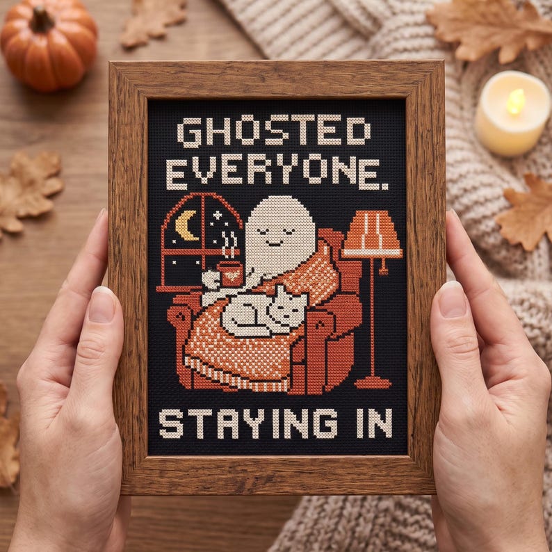 May include: A framed cross-stitch embroidery on black fabric featuring a white ghost sitting in an orange armchair with a cat, holding a mug, with a window and lamp. The text reads GHOSTED EVERYONE. STAYING IN in white block letters.