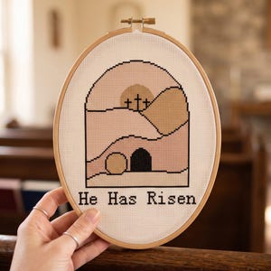 K&ouml;nnte beinhalten: Ein ovaler Stickrahmen aus Holz mit wei&szlig;em Aida-Stoff und einem Kreuzstichmotiv. Das Muster zeigt drei schwarze Kreuze auf einem H&uuml;gel, eine kreisf&ouml;rmige Sonne und ein offenes Grab in Beige- und Rosat&ouml;nen. Der Text "He Has Risen" ist in Schwarz gestickt.