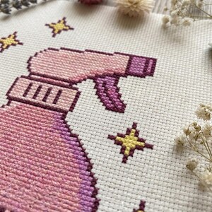Peut inclure: Broderie au point de croix repr&eacute;sentant un vaporisateur rose et violet avec des &eacute;toiles jaunes. Le motif est sur un tissu blanc, avec une vue rapproch&eacute;e montrant les points de couture d&eacute;taill&eacute;s. L'&oelig;uvre est entour&eacute;e de fleurs s&eacute;ch&eacute;es.
