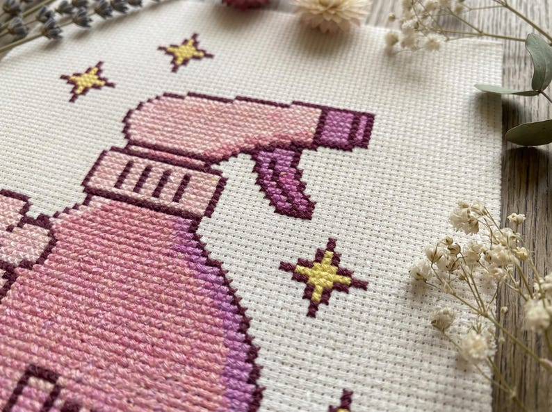 Peut inclure: Broderie au point de croix repr&eacute;sentant un vaporisateur rose et violet avec des &eacute;toiles jaunes. Le motif est sur un tissu blanc, avec une vue rapproch&eacute;e montrant les points de couture d&eacute;taill&eacute;s. L'&oelig;uvre est entour&eacute;e de fleurs s&eacute;ch&eacute;es.