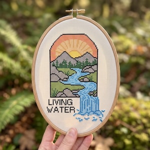 K&ouml;nnte beinhalten: Ein ovaler Stickrahmen aus Holz mit einem Kreuzstichmotiv, das eine Landschaft mit Sonnenuntergang, Bergen und einem blauen Wasserfall zeigt. Der Text LIVING WATER ist in schwarzen Buchstaben unten links auf dem wei&szlig;en Stoff gestickt.
