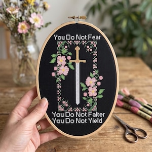 May include: An oval wooden embroidery hoop containing black fabric with a cross-stitch design. The design features a central gold sword surrounded by pink flowers and green leaves, with the text "You Do Not Fear, You Do Not Falter, You Do Not Yield" stitched in white.
