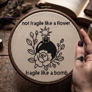 K&ouml;nnte beinhalten: Ein runder Stickrahmen aus Holz mit einem Kreuzstichmotiv auf cremefarbenem Stoff. Das Design zeigt eine schwarze Bombe und eine Rose mit dem Text "not fragile like a flower" oben und "fragile like a bomb" am unteren Rand.