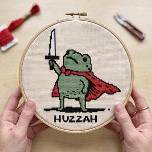 May include: A circular wooden embroidery hoop containing a cross-stitch design of a green frog wearing a red cape and holding a silver sword aloft. The word HUZZAH is stitched in black capital letters at the bottom of the cream-coloured fabric.