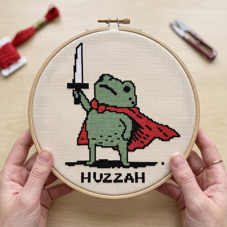 May include: A circular wooden embroidery hoop containing a cross-stitch design of a green frog wearing a red cape and holding a silver sword aloft. The word HUZZAH is stitched in black capital letters at the bottom of the cream-coloured fabric.