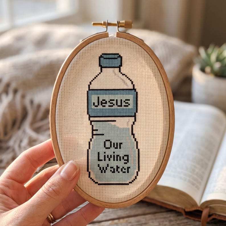 Puede incluir: Un bastidor de bordado ovalado de madera que contiene un dise&ntilde;o de punto de cruz de una botella de agua. La botella est&aacute; rellena con hilo azul claro y presenta el texto negro "Jesus" en la etiqueta y "Our Living Water" en la parte inferior.