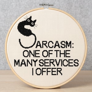 May include: A circular wooden embroidery hoop containing a piece of off-white Aida fabric. The fabric features a black cross-stitch design of a cat shaped like the letter S, followed by the text: SARCASM: ONE OF THE MANY SERVICES I OFFER.