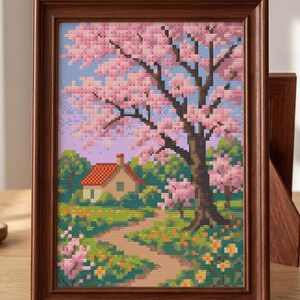 Puede incluir: Paisaje en punto de cruz enmarcado que presenta una casa con tejado rojo, un camino y un gran &aacute;rbol con flores rosas. La obra de arte est&aacute; enmarcada en un marco de madera, mostrando una serena escena primaveral con colores vibrantes.