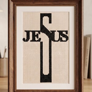 Puede incluir: Una pieza de bordado en punto de cruz con la palabra JESUS en hilo negro, donde la letra S forma la barra vertical de una cruz. La obra est&aacute; expuesta en un marco de madera marr&oacute;n oscuro con un paspart&uacute; blanco sobre un fondo neutro.