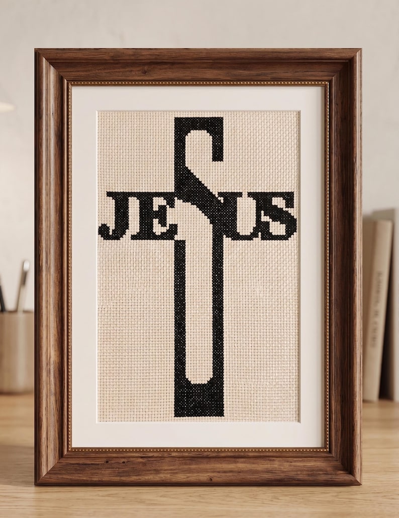Puede incluir: Una pieza de bordado en punto de cruz con la palabra JESUS en hilo negro, donde la letra S forma la barra vertical de una cruz. La obra est&aacute; expuesta en un marco de madera marr&oacute;n oscuro con un paspart&uacute; blanco sobre un fondo neutro.