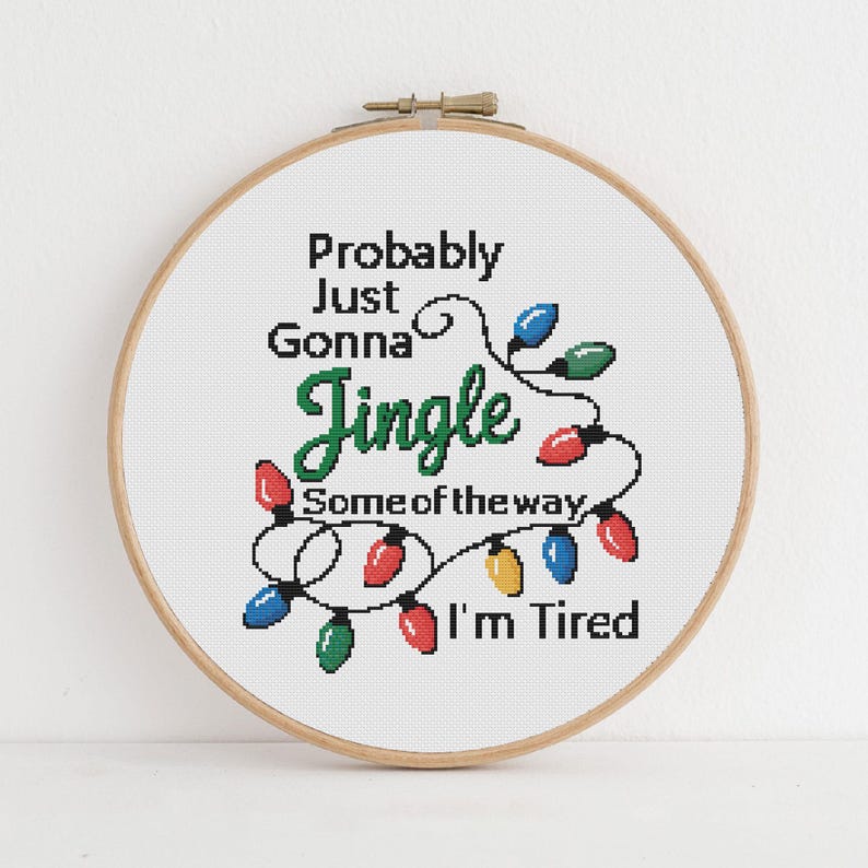 Może przedstawiać: Obręcz do haftu krzyżykowego z napisem "Probably Just Gonna Jingle Some of the way, I'm Tired." Projekt zawiera kolorowe lampki choinkowe w kolorze czerwonym, zielonym, niebieskim i ż&oacute;łtym. Obręcz jest drewniana.