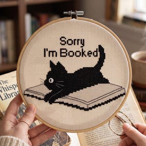 May include: A circular wooden embroidery hoop containing a cross-stitch design on cream fabric. The design features a black cat lying on an open book with the black text Sorry I'm Booked stitched above the cat. The hoop has a metal tension screw.