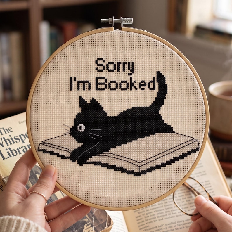 May include: A circular wooden embroidery hoop containing a cross-stitch design on cream fabric. The design features a black cat lying on an open book with the black text Sorry I'm Booked stitched above the cat. The hoop has a metal tension screw.