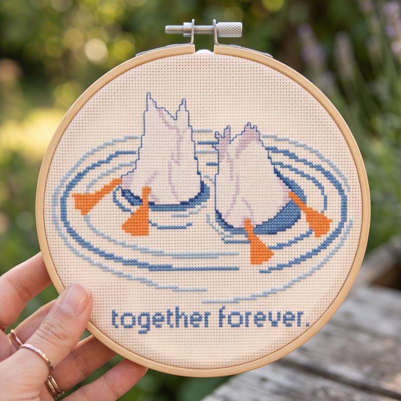 Puede incluir: Un bastidor de bordado circular de madera que contiene un dise&ntilde;o de punto de cruz de dos patos buceando en el agua con sus patas naranjas visibles sobre la superficie. El texto "together forever." est&aacute; bordado en azul en la parte inferior.