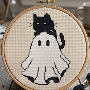 May include: A cross-stitch pattern featuring a black cat perched atop a white ghost. The design is set within a wooden embroidery hoop. The text "CROSS STITCH PATTERN - INSTANT DOWNLOAD" is at the top. Black thread and scissors are visible.