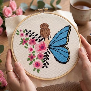 Peut inclure: Un projet de broderie au point de croix dans un tambour en bois circulaire repr&eacute;sentant une personne avec un chignon brun, une grande aile de papillon bleue et un bouquet de fleurs roses avec des feuilles vertes sur un tissu cr&egrave;me.