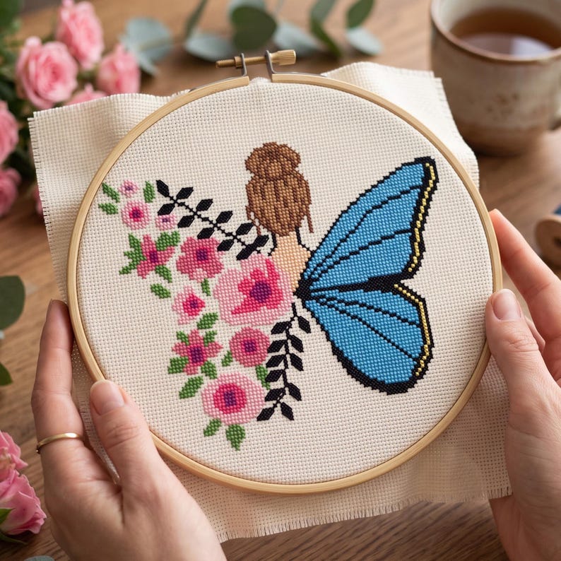 Peut inclure: Un projet de broderie au point de croix dans un tambour en bois circulaire repr&eacute;sentant une personne avec un chignon brun, une grande aile de papillon bleue et un bouquet de fleurs roses avec des feuilles vertes sur un tissu cr&egrave;me.