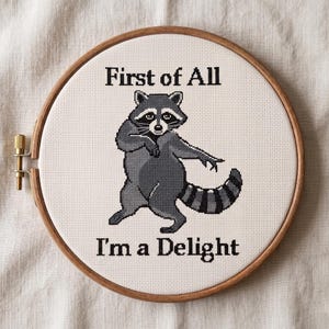 May include: A cross-stitch embroidery in a wooden hoop shows a raccoon with the text "First of All I'm a Delight." The raccoon is in shades of grey and black, with a striped tail and a playful pose. The background is a light beige fabric.