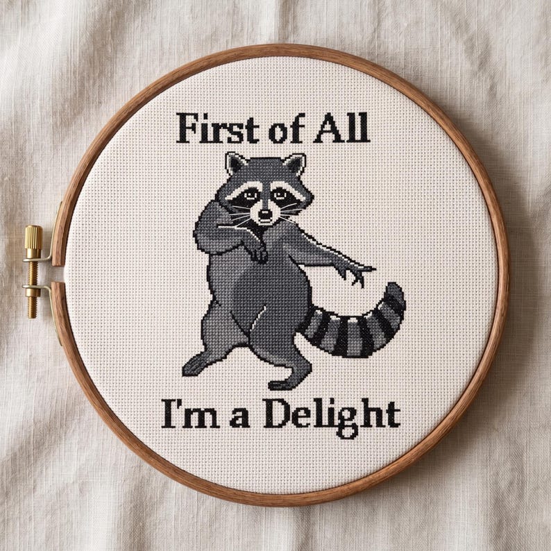 May include: A cross-stitch embroidery in a wooden hoop shows a raccoon with the text "First of All I'm a Delight." The raccoon is in shades of grey and black, with a striped tail and a playful pose. The background is a light beige fabric.