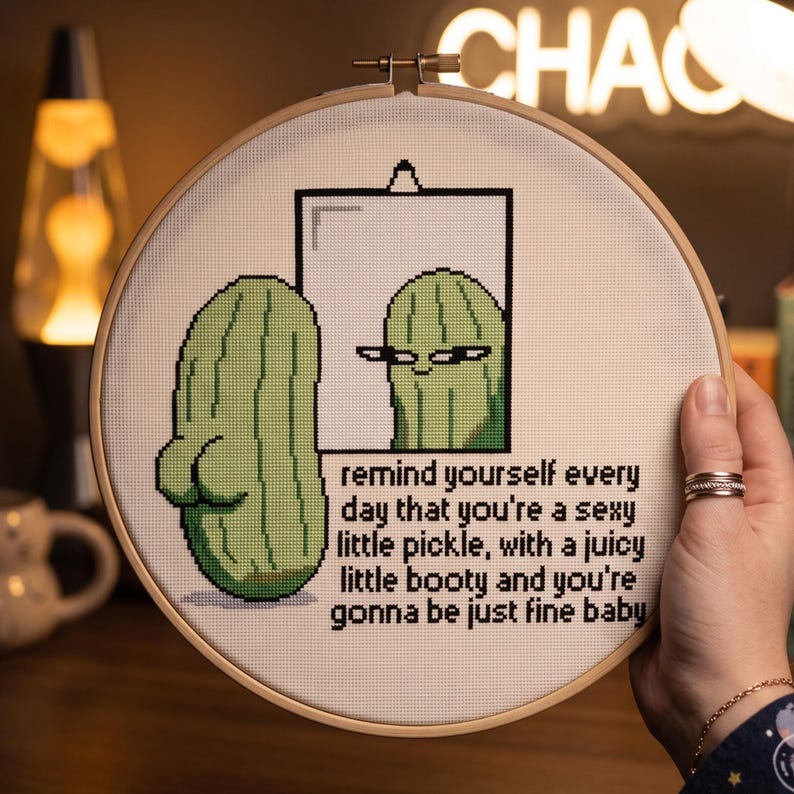 May include: A circular wooden embroidery hoop containing a cross-stitch design of a green pickle looking into a mirror. The text reads: remind yourself every day that you're a sexy little pickle, with a juicy little booty and you're gonna be just fine baby.