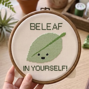 Puede incluir: Un bastidor de bordado circular de madera que contiene un dise&ntilde;o de punto de cruz sobre tela blanca. El dise&ntilde;o presenta una hoja de color verde claro con una cara negra sencilla y el texto verde oscuro BELEAF IN YOURSELF! centrado.