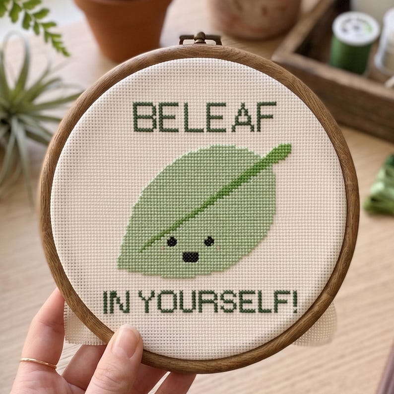 Puede incluir: Un bastidor de bordado circular de madera que contiene un dise&ntilde;o de punto de cruz sobre tela blanca. El dise&ntilde;o presenta una hoja de color verde claro con una cara negra sencilla y el texto verde oscuro BELEAF IN YOURSELF! centrado.