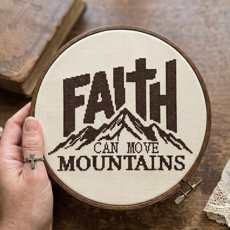 May include: A circular wooden embroidery hoop containing a piece of off-white fabric with dark brown cross-stitched embroidery. The design features the word FAITH with a cross integrated into the letter T, a mountain range, and the text CAN MOVE MOUNTAINS.