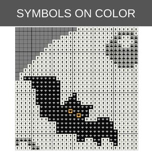 Pu&ograve; includere: Uno schema per punto croce intitolato SYMBOLS ON COLOR che presenta una griglia con quadrati neri, bianchi e grigi. Il disegno mostra la sagoma di un pipistrello nero con due occhi arancioni a forma di cuore, circondato da simboli di frecce e tazzine.