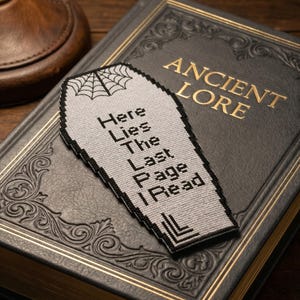 May include: A gray coffin-shaped bookmark with black trim and a spiderweb design at the top. The text "Here Lies The Last Page I Read" is stitched on the bookmark. The bookmark rests on a dark gray book with gold lettering that reads "Ancient Lore".
