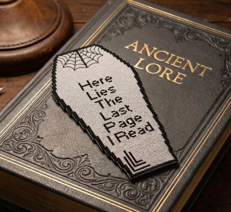May include: A gray coffin-shaped bookmark with black trim and a spiderweb design at the top. The text "Here Lies The Last Page I Read" is stitched on the bookmark. The bookmark rests on a dark gray book with gold lettering that reads "Ancient Lore".