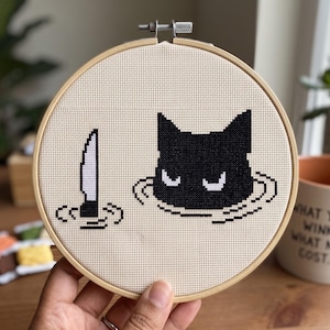 K&ouml;nnte beinhalten: Ein runder h&ouml;lzerner Stickrahmen mit einem Kreuzstichmotiv auf beigem Stoff. Das Design zeigt einen schwarzen Katzenkopf mit wei&szlig;en Augen, der aus dem Wasser ragt, neben einem vertikalen Messer, das ebenfalls aus dem Wasser ragt.
