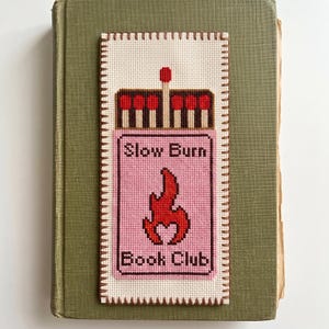 Puede incluir: Un marcap&aacute;ginas rectangular de punto de cruz con un dise&ntilde;o de caja de cerillas sobre fondo rosa, un gr&aacute;fico de llama roja y el texto "Slow Burn Book Club" en marr&oacute;n oscuro. El marcap&aacute;ginas tiene un borde crema con costuras marrones.