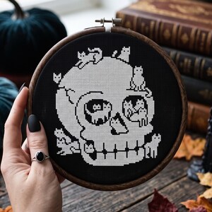 May include: A circular wooden embroidery hoop containing a black fabric piece with a white cross-stitch design of a human skull. The skull is composed of multiple small cat figures, with cats positioned on the cranium, inside the eye sockets, and along the jawline.