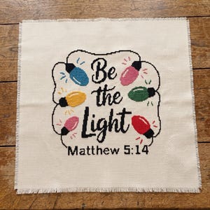 Może przedstawiać: Kwadratowy kawałek kremowej tkaniny Aida z haftem krzyżykowym przedstawiającym czarny napis "Be the Light Matthew 5:14" otoczony kolorowymi niebieskimi, ż&oacute;łtymi, r&oacute;żowymi, zielonymi i czerwonymi żar&oacute;wkami świątecznymi z małymi promienistymi ściegami.