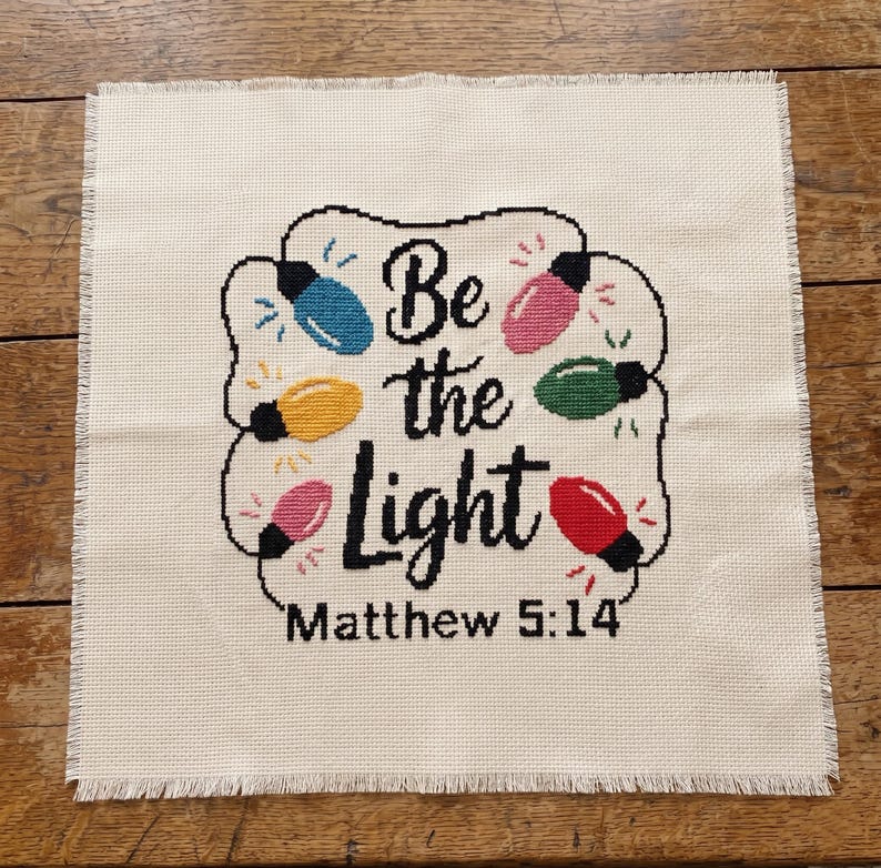 Może przedstawiać: Kwadratowy kawałek kremowej tkaniny Aida z haftem krzyżykowym przedstawiającym czarny napis "Be the Light Matthew 5:14" otoczony kolorowymi niebieskimi, ż&oacute;łtymi, r&oacute;żowymi, zielonymi i czerwonymi żar&oacute;wkami świątecznymi z małymi promienistymi ściegami.