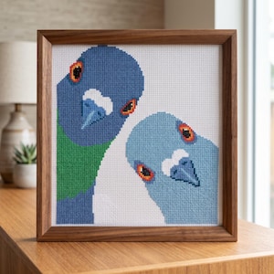 May include: A framed cross-stitch artwork featuring two pigeons. The birds have blue heads, orange eyes, and white beaks. One pigeon has a green neck. The artwork is set against a white background and framed in a brown wooden frame.