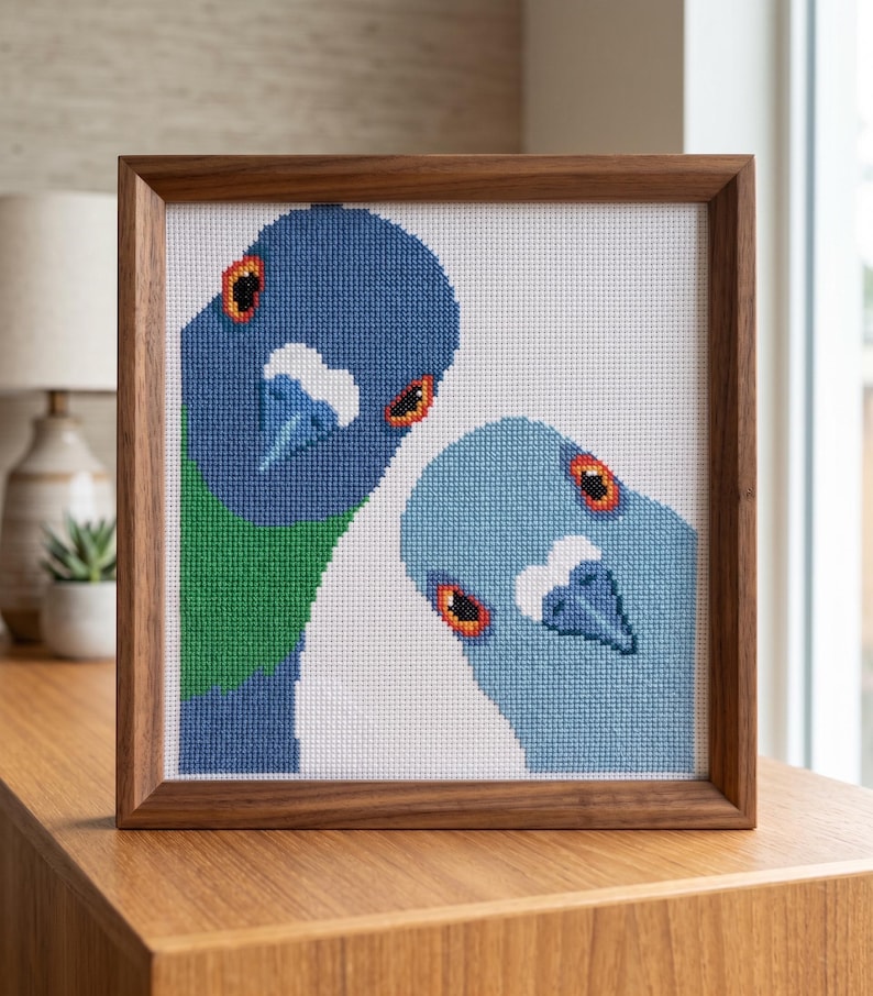 May include: A square cross-stitch embroidery featuring two pigeons, one dark blue with green accents and one light blue, set against a white background. The artwork is encased in a simple, medium-toned wooden frame with a clean, square profile.