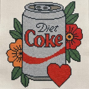 May include: A cross-stitch embroidery on cream fabric featuring a silver soda can with the text Diet Coke in red and black. The can is surrounded by orange and red flowers with green leaves and a red heart positioned at the bottom right corner.