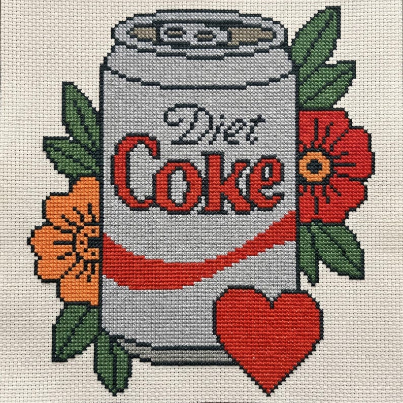 May include: A cross-stitch embroidery on cream fabric featuring a silver soda can with the text Diet Coke in red and black. The can is surrounded by orange and red flowers with green leaves and a red heart positioned at the bottom right corner.