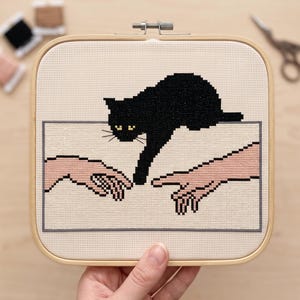 Pode incluir: Um bastidor de bordado quadrado de madeira contendo um design em ponto cruz sobre tecido creme. O padr&atilde;o apresenta duas m&atilde;os de tons claros estendendo-se uma para a outra, com um gato preto acima, esticando uma pata em dire&ccedil;&atilde;o aos dedos.